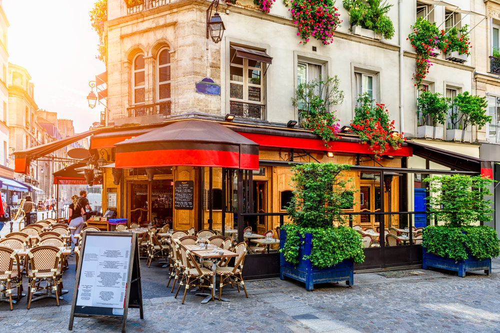 Pretty building with red awning on cobblestone street with menu sign and lush greenery and matching beige and brown tables and chairs.