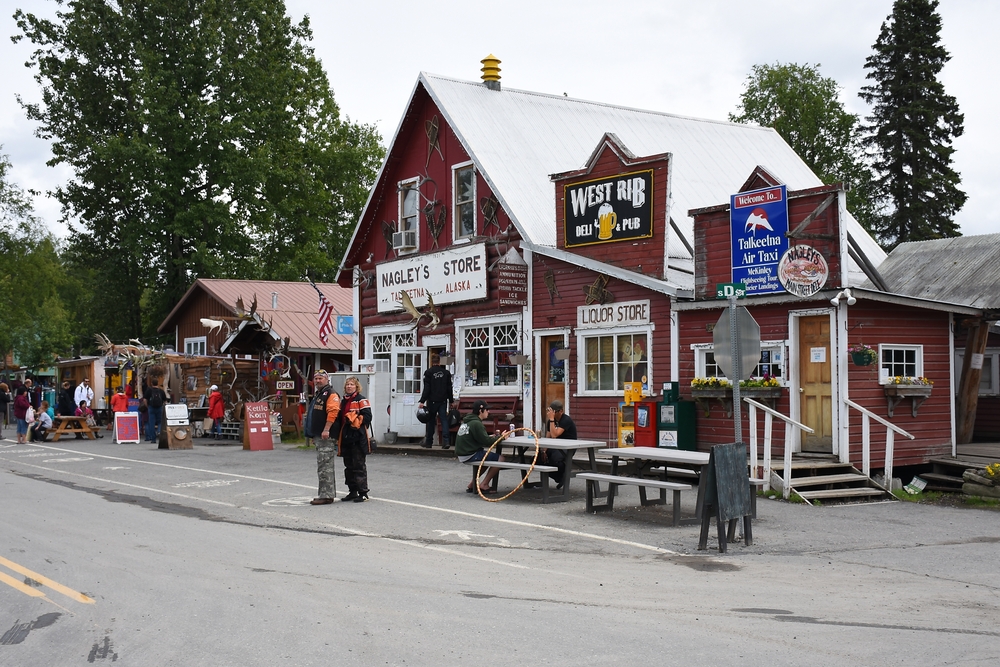 Front of a red store in Talkeetna with many signs on it and people walking around.