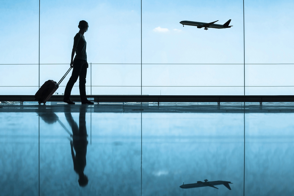 Woman walking past wall of glass windows with plane taking off in background, pulling carry-on suitcase. 
How to pack lightly using only  a carry-on.