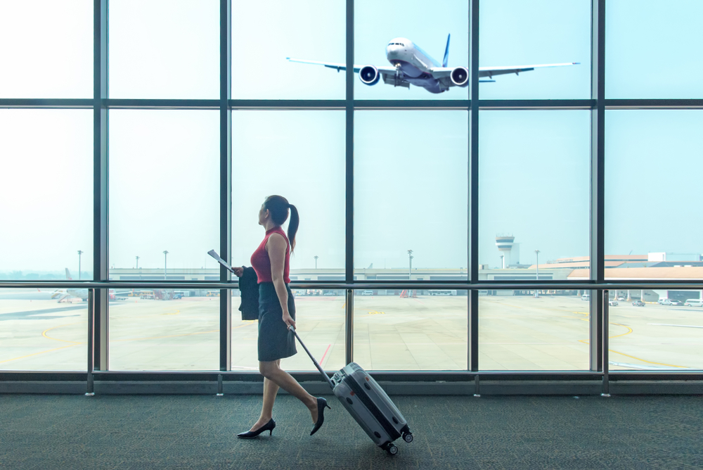 Woman walking next to wall of glass windows with airplane taking off, carrying papers pulling silver carryon suitcase. How to pack lightly in a carryon.