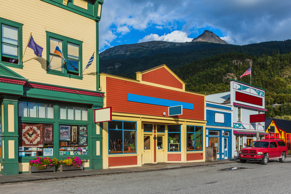 Colorful storefront towns in Alaska with red SUV parked in front and green mountain in background.