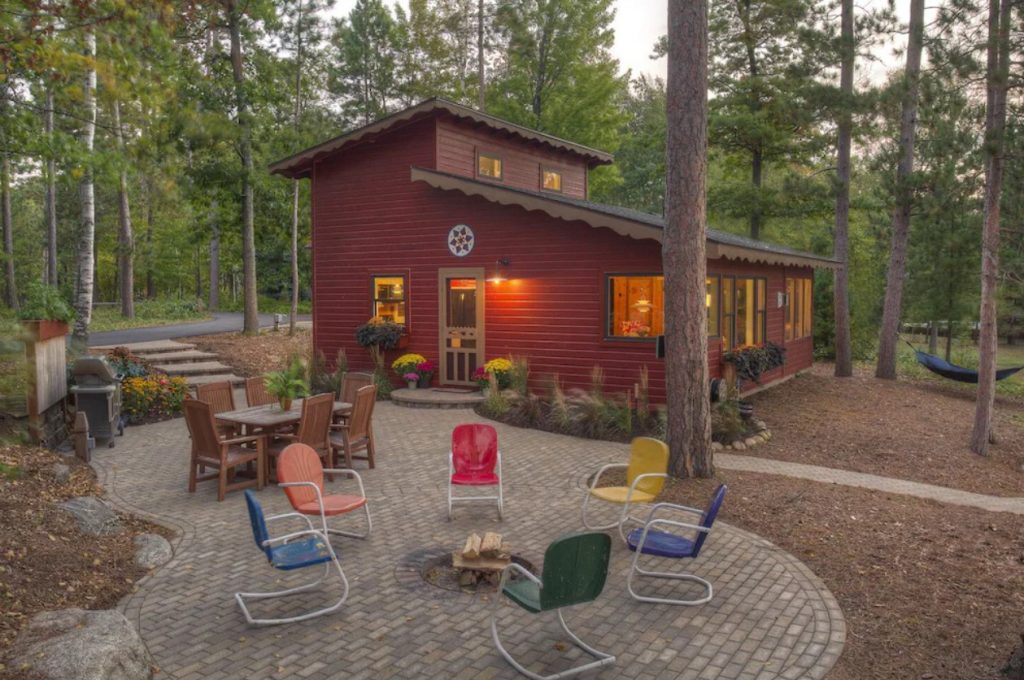 The exterior of a charming red cabin in the woods, one of the best cabins in the Midwest. The cabin has scalloped brown trim along the roof line, lots of windows, and a circular plaque with a painted flower on it. There is also a stone patio with multi colored chairs around a fire pit, a dining set, and a grill. 
