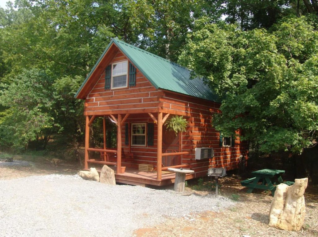 A small reddish orange log cabin with a green roof. It has a small front porch with a porch swing and next to the cabin is a green picnic table. There is a pebbled area in front of the cabin and it is surrounded with trees. One of the best cabins in the Midwest. 