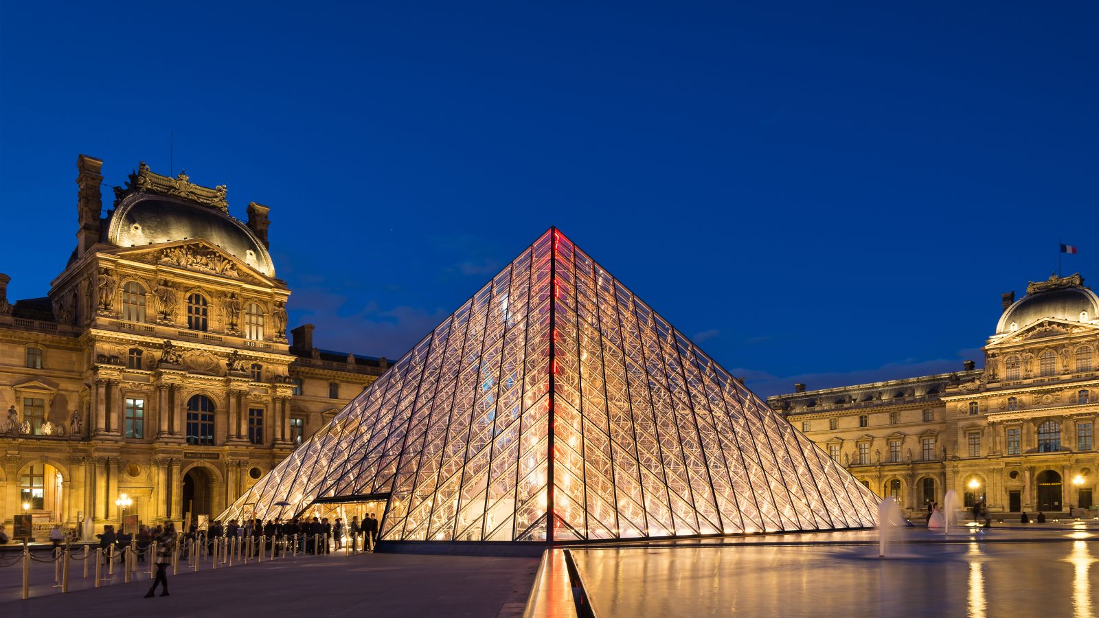 Illuminated glass pyramid with ornate building in background.