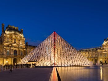Illuminated glass pyramid with ornate building in background.