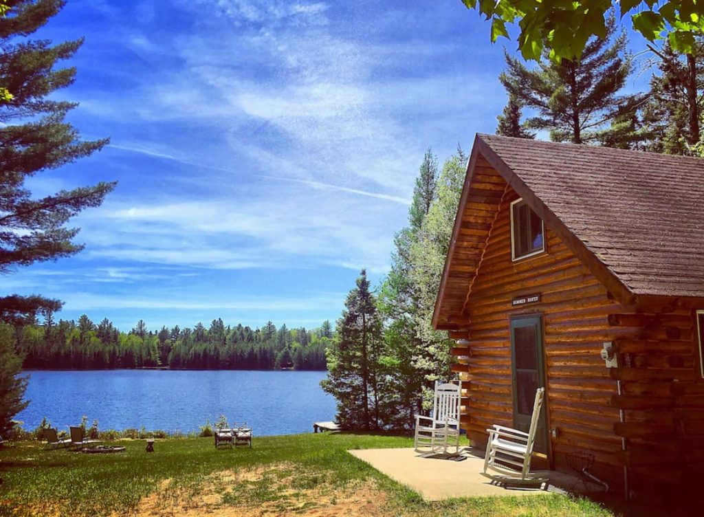 The exterior of the front of a classic log cabin with a small patio in front of it. The patio has two white rocking chairs. Behind the cabin there is a grassy shore that leads to a lake with trees all around it. 