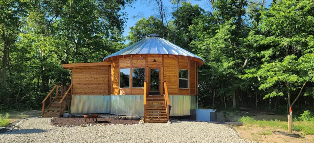 The exterior of one of the coolest cabins in the Midwest. It is circular shaped, like a yurt, but made out of wooden logs. There is a small staircase leading to the front door, an aluminum roof, and it is surrounded by trees. 
