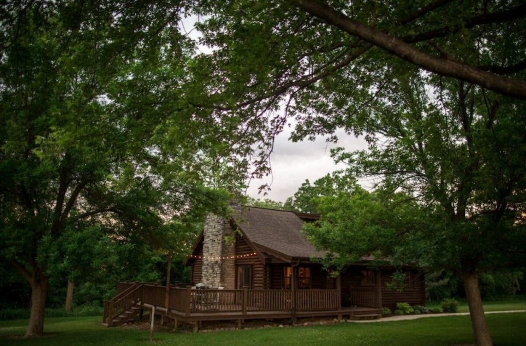 A log cabin with a large porch, a stone fireplace, and twinkle lights hanging over the porch. It is surrounded by a green lawn and lush green trees. 