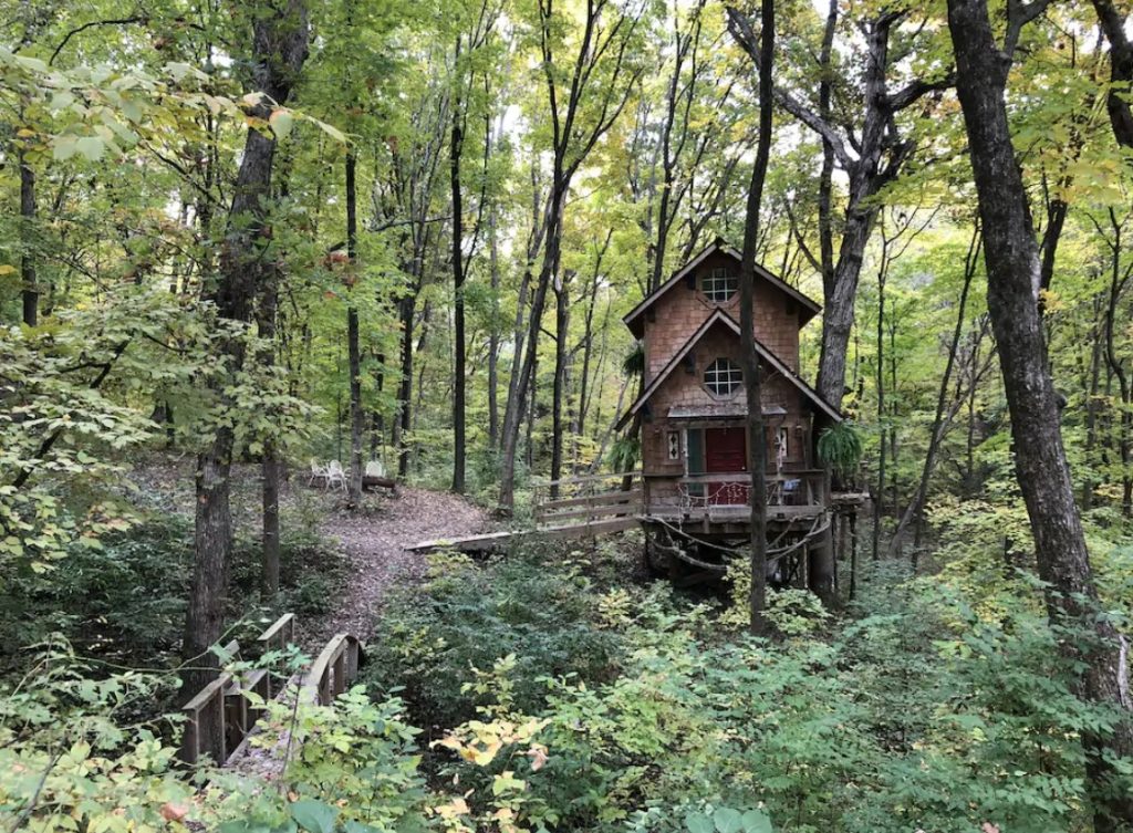 A cabin on the side of a hill surrounded by a dense forest. It has two levels each with a window in the peak. You can see a front porch, the front door, and twinkle lights hanging on the railing. Near the ramp to get to the cabin you can see white chairs and a pathway.