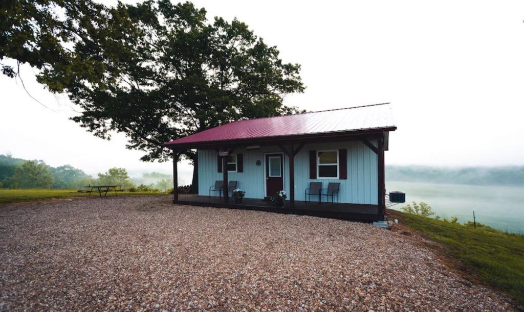 The exterior of a white metal cabin with a red roof and a small front porch with chairs on it. In front of the cabin is a large pebble area and you can see a picnic table off to the side. Behind the cabin you can see a grassy lawn, large trees, and fog on a field. One of the best cabins in the Midwest. 