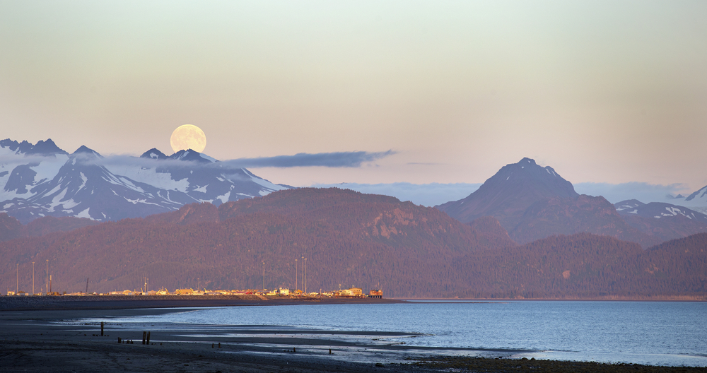 Moon in sky over snow-capped mountains, with town and beach in foreground.