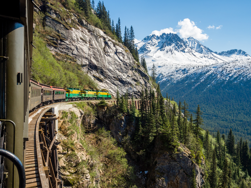 Colorful train on side of mountain with snow-capped mountains in background. Evergreen trees in foreground. Traveling to Alaska