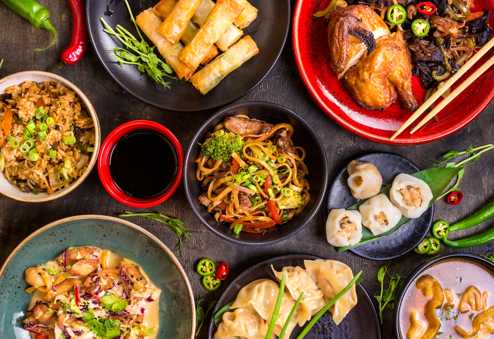 An overhead image of red and black plates full of Chinese food like the food you can get at one of the best restaurants in Philadelphia. There is rice, noodles, dumplings, spring rolls, chicken, peppers, soy sauce, and chopsticks. 