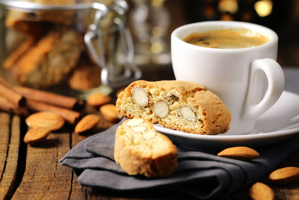 A almond scone sitting on black napkin and on a saucer. There is a coffee cup on the saucer as well and it is filled with a coffee that has a frothy top. The cup and saucer are white. 