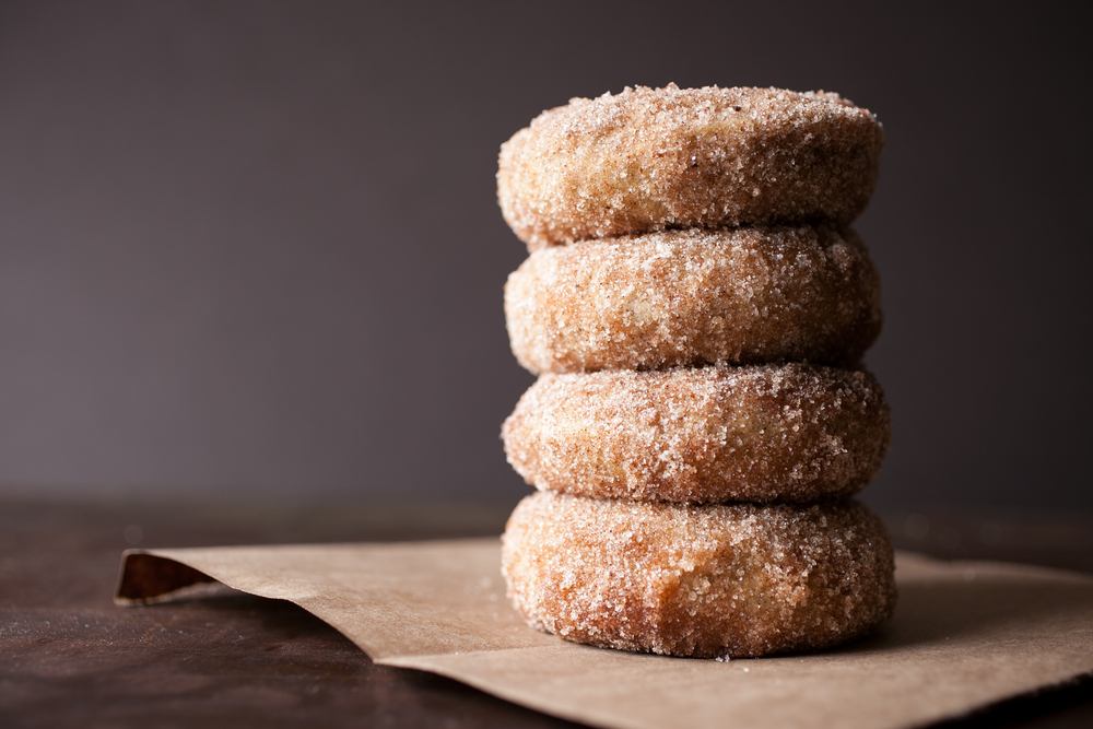 A stack of four donuts covered in cinnamon sugar. They are on top of a piece of brown paper on a wooden table. 