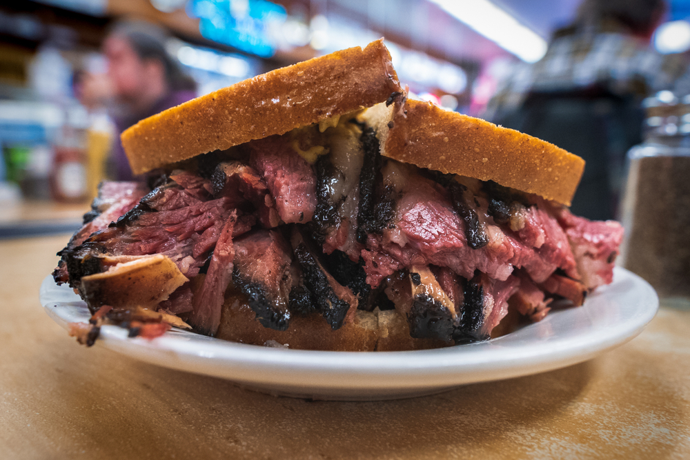 A large sandwich on a small white plate. It is overflowing with beef with charred tips and you can see some cheese. It is on a slice of what looks like white bread. 