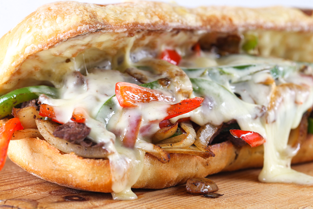 A close up of a cheesesteak like one you can find at one of the best restaurants in Philadelphia. It has beef, onions, peppers, mushrooms, cheese, and is on a toasted bun.