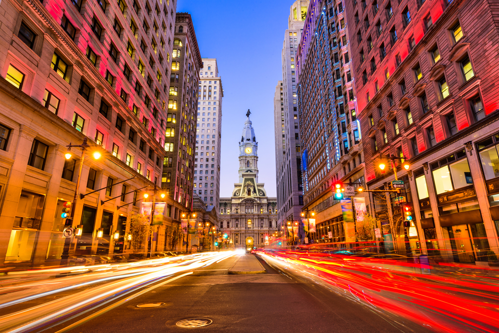 A main street in Philadelphia. It is a long exposure photograph with cars making light trails on the street. It is twilight and the buildings are lit up.
