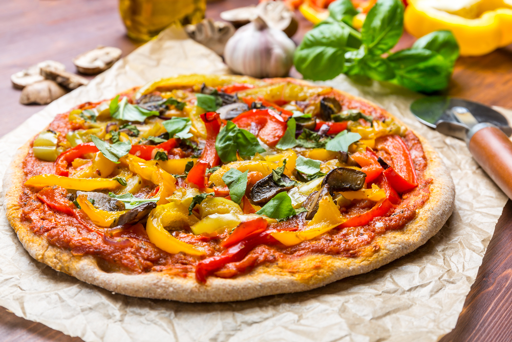 A large pizza on a piece of crinkled wax paper. The pizza has peppers, onions, sauce, basil, and other spices on it. In the background you can see some vegetables and a pizza cutter. 