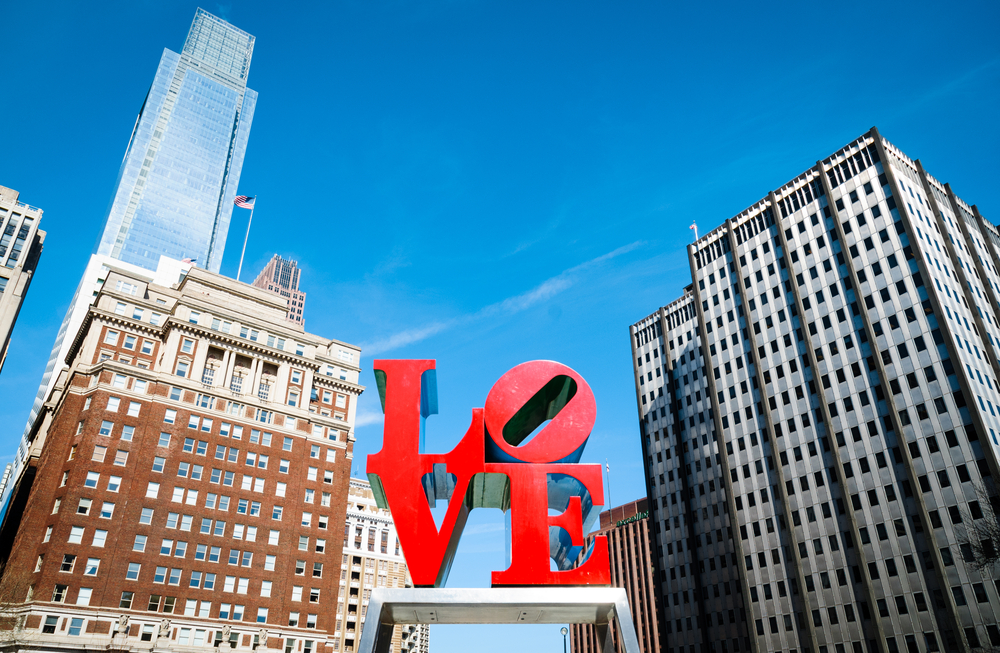 Looking up at the famous LOVE sculpture in LOVE Park. It is a bright red statue with the LO stacked on top of VE. In the background you can see part of the Philly skyline. 