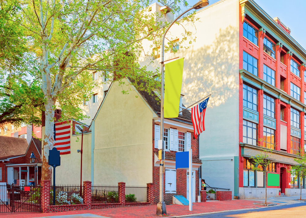 The exterior of a small house that is believed to be where Betsy Ross stitched the first American Flag. It is a very small brick building with white windows and doors and a small brick courtyard. There is a large tree in the courtyard and modern buildings around it. 