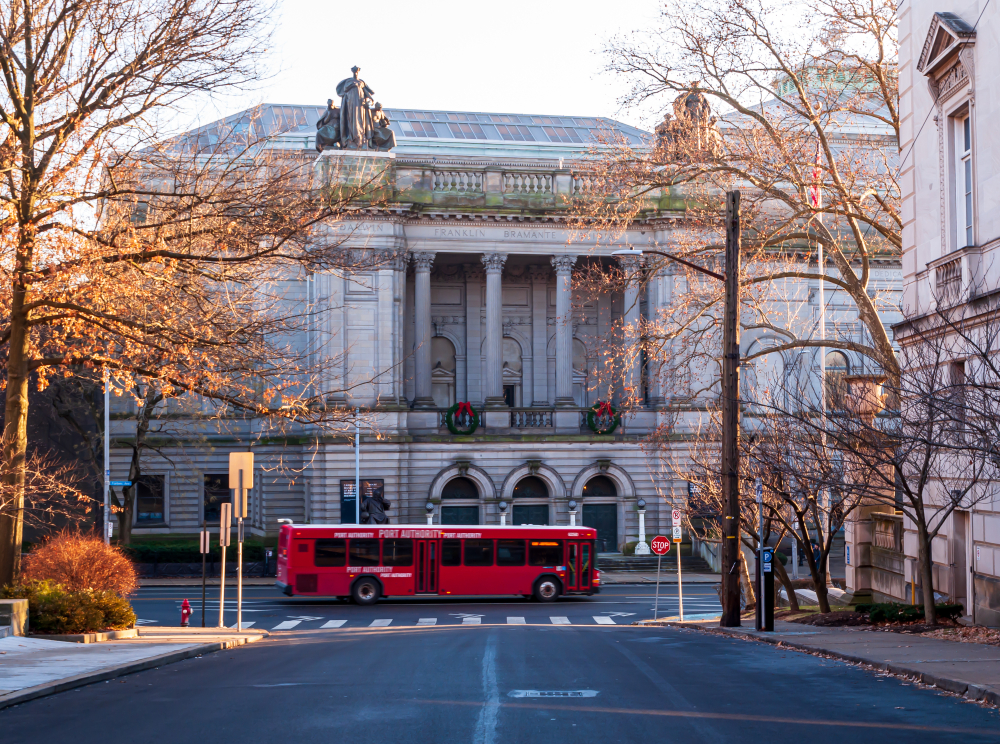 The exterior of a grand building that is the Carnegie Museum of Natural History. In front of it is a red bus and it is surround with trees with no leaves. 