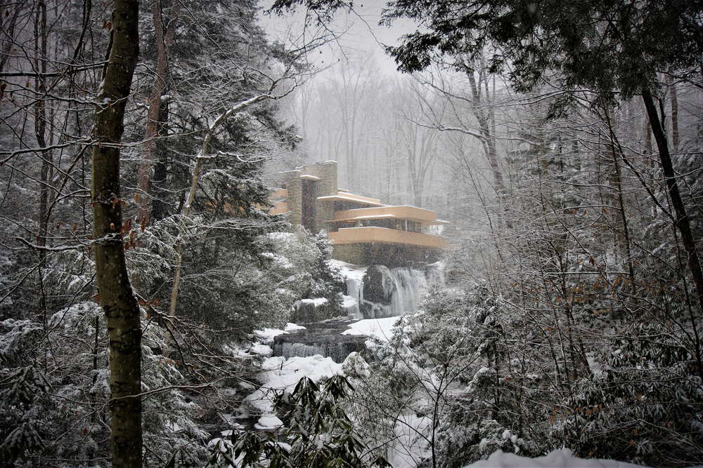 A view of Frank Lloyd Wright's Fallingwater in the snow. You can see the waterfall frozen underneath the house and flowing into the semi frozen river. The house, trees, and rocks in the river are covered in snow. The sky is gray. 