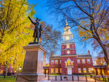 A large brick building that is the Independence Hall, one of the best things to do in Philadelphia. In front of it are large trees, and a statue of a founding father on a marble base.