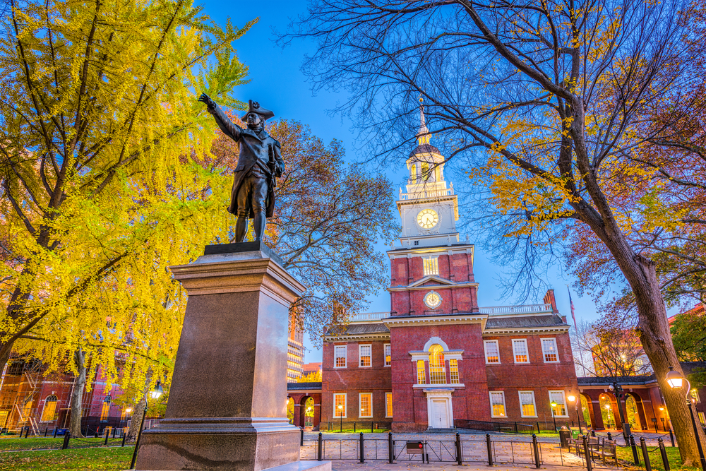 A large brick building that is the Independence Hall, one of the best things to do in Philadelphia. In front of it are large trees, and a statue of a founding father on a marble base.