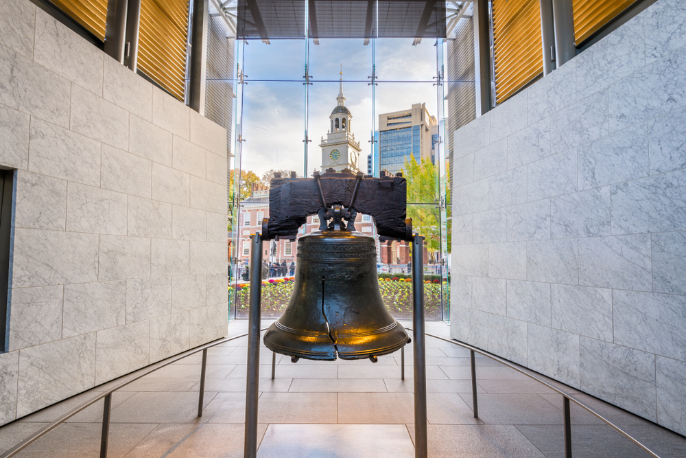 The Liberty Bell on display inside a building in Philadelphia. It is surrounded by marble walls and there is a glass window that looks out onto Independence Plaza. 