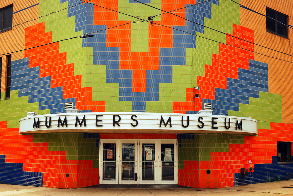 The exterior of the Mummer's Museum in Philly. It is an Mid Century Modern style building that used to be an old movie theater. There is a large mosaic on the front made of red, green, and blue tiles in a chevron pattern up the entire front where the entrance is.