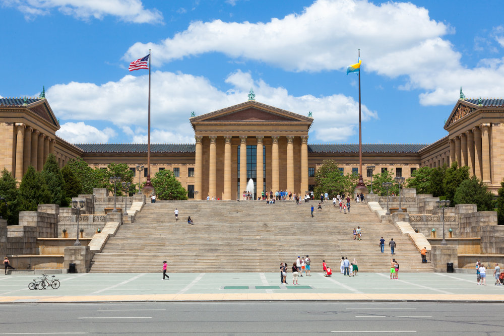 The front exterior of a large ornate Greek style building. It is the Philadelphia Museum of Art. There are large rows of steps in front of it and you can see people walking around. 