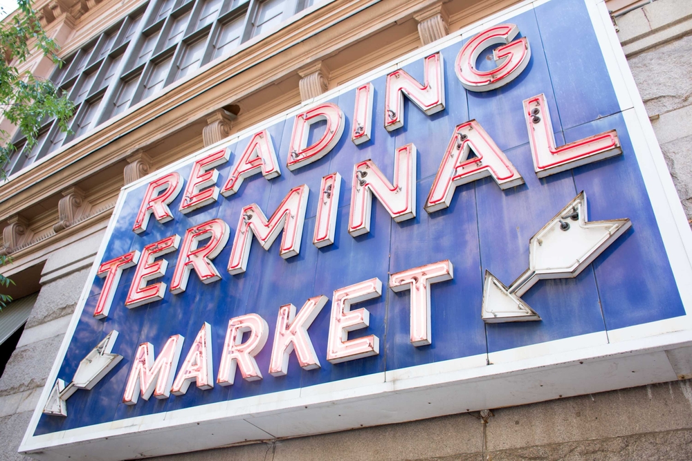 A large blue sign with a red neon sign that reads 'Reading Terminal Market'. There are two white neon arrows pointing down. The sign is on the side of a building. 