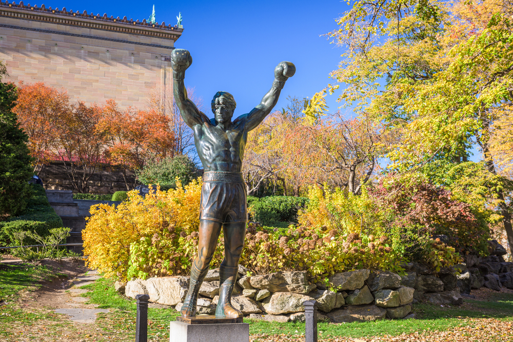 A bronze statue of Rocky Balboa, a character from the Rocky movies. It is one of the best things to do in Philadelphia. Behind the statue you can see lush gardens with grasses, shrubs, trees, and other plants. 