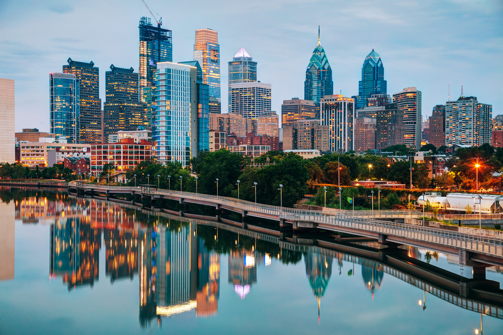 The Philadelphia skyline at twilight. You can see the Schuylkill River next to the city. The city is all lit up and you can see the reflection in the river.