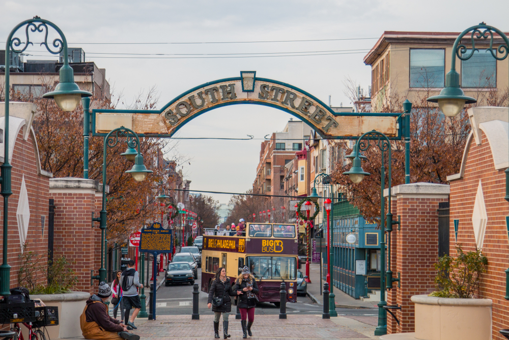 The entrance of South Street in Philly. You can see a bus, people walking, and shops down the street. The street is decorated for the holidays so there are wreaths and bright red light poles with garland and ornaments on them. 