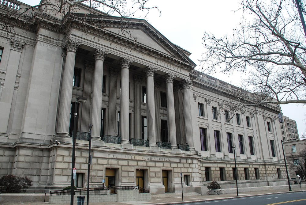 The exterior of a large gray Greek-colonial building. It is very large with several floors and sits right on the side of the street. 