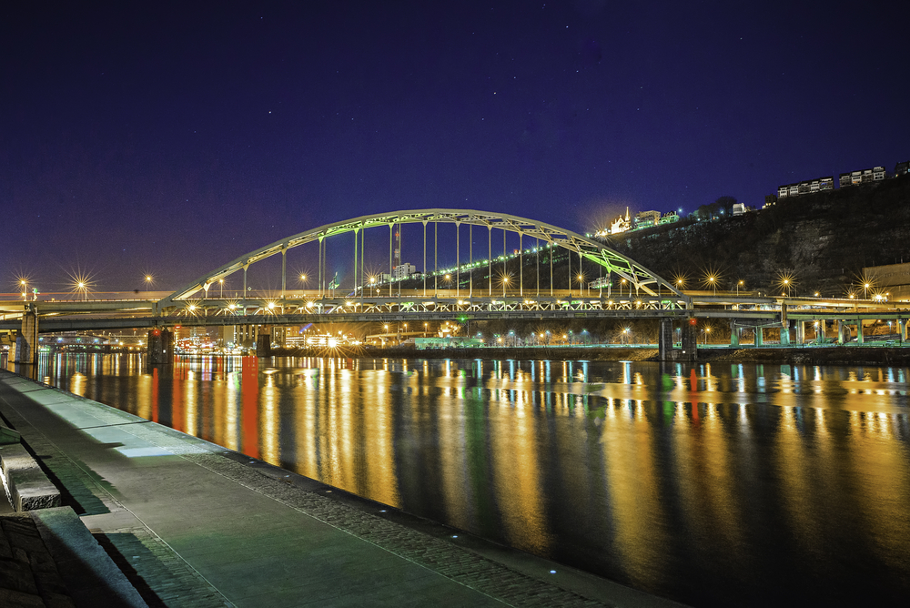 The view of the Allegheny River at night along the river bank. You can see lights reflecting in the water, a large bridge that is lit up, and buildings on a hill in the distance. 