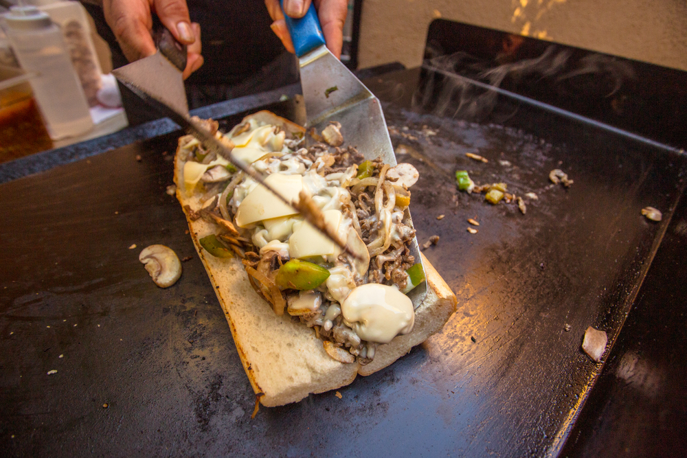 A person using spatulas to place toppings onto a bun. The toppings are a cheesesteak, so there is thinly sliced beef, onion, mushrooms, peppers, and provolone cheese. It's one of the best things to do in Philadelphia.