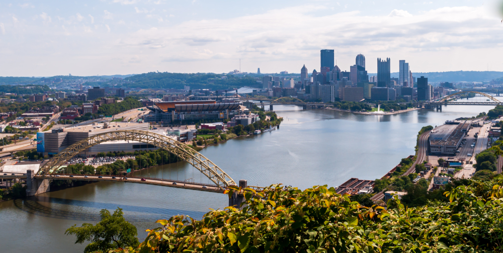 A scenic view of the Pittsburgh skyline, the Allegheny River, and a rounded bridge over the river. 