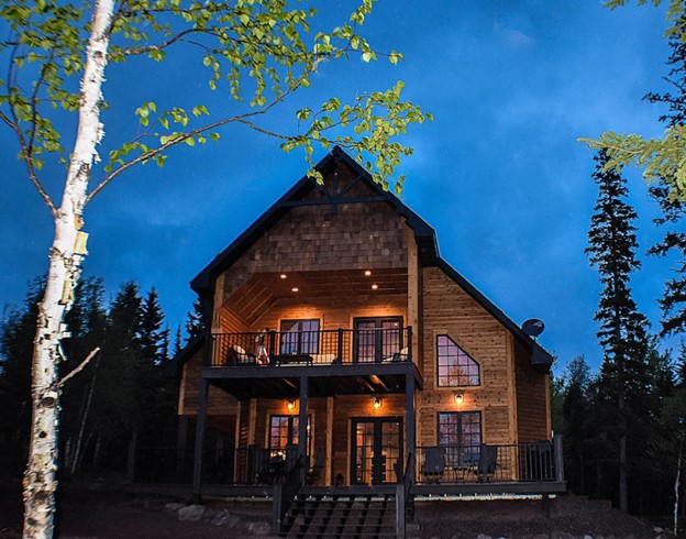 Beautiful cabin with large windows with large front porch and balcony, with romantic lights against blue night sky.