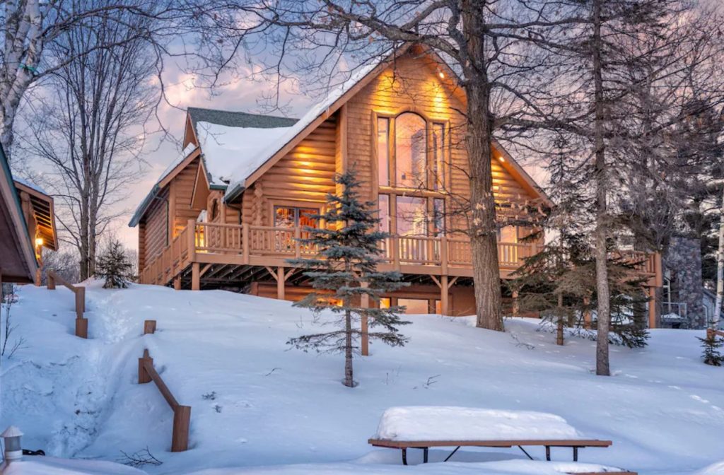 The exterior of a large and luxurious cabins in Minnesota. There is a large window in the front of it and a wrap around porch. The ground and the cabin are covered in snow. 