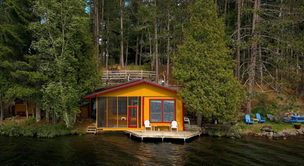 A orangish yellow cabin with red accents that sits right on a lake. Behind it you can see a dense forest. One of the best cabins in Minnesota. 