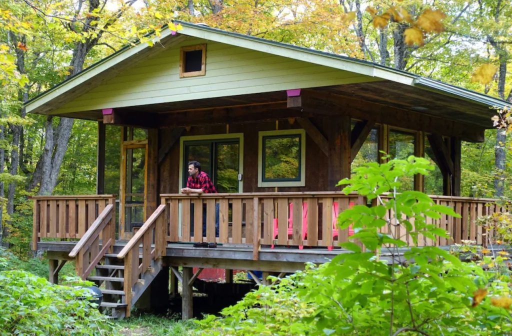 A man standing on a wrap around porch attached to a small cabin. It is surrounded by maple trees and almost every wall is windows.
