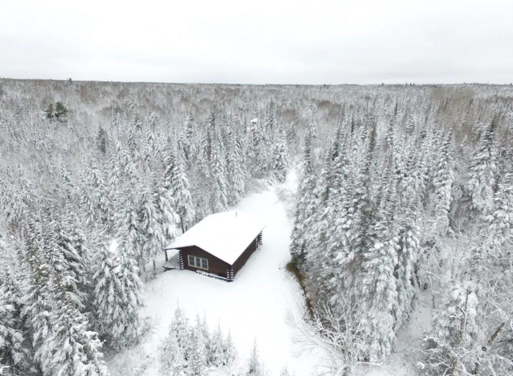 A log cabin covered in snow. It is surrounded by a dense forest that is also covered in snow. 
