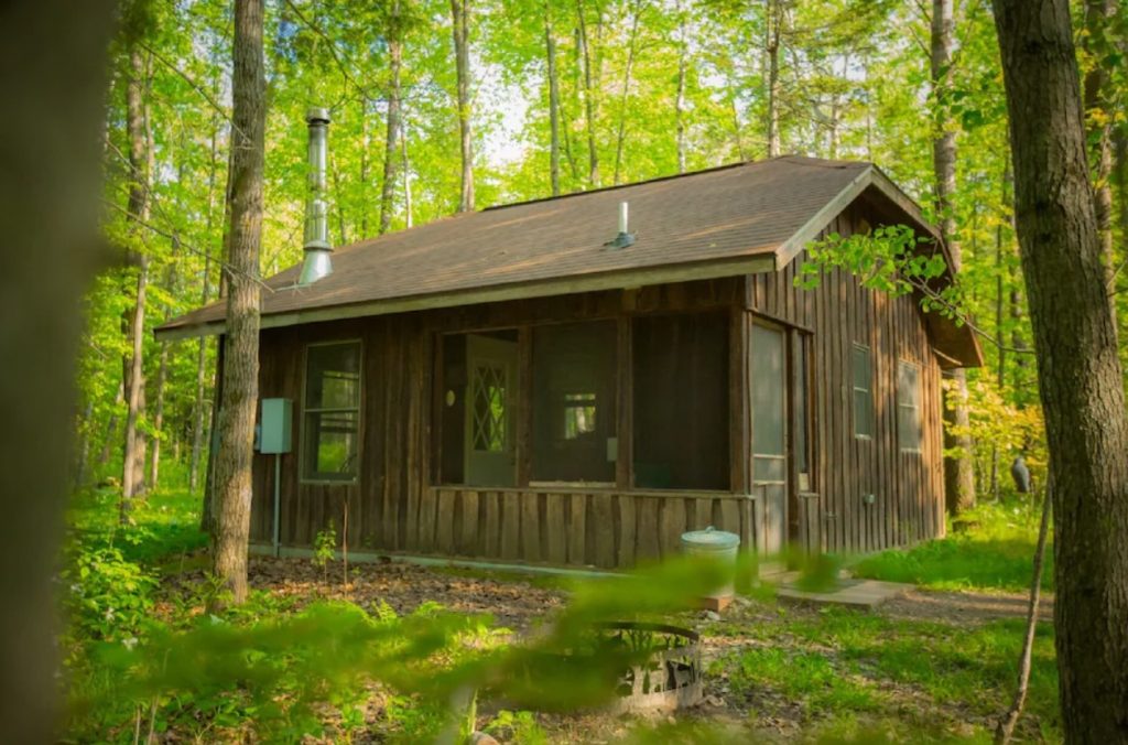 A dark wood paneled cabin in the middle of a forest in Minnesota. There is a screened in porch and windows on the side of the cabin. 