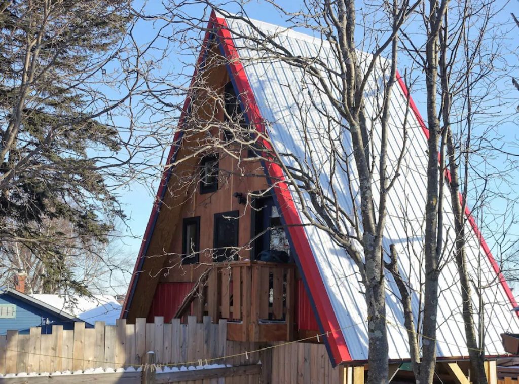 The exterior of a classic A-frame style cabin with red trim on the roof. You can see windows and a small deck. It is surrounded by trees. 