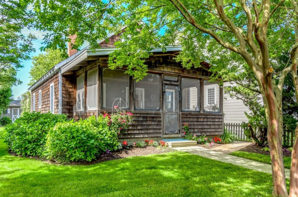 The front of a weathered wooden cottage. It is surrounded by a green yard, shrubs, trees, and a small flower garden with pink and red flowers. One of the best Delaware Airbnbs. 