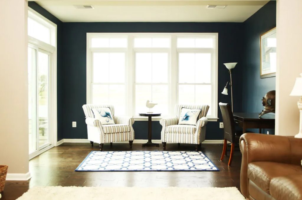 A living room with dark blue walls, stripped chairs, and a brown leather sofa. There several windows in the rooms. 