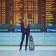 Woman with long brown hair wearing black suit with luggage looking at airplane departures board